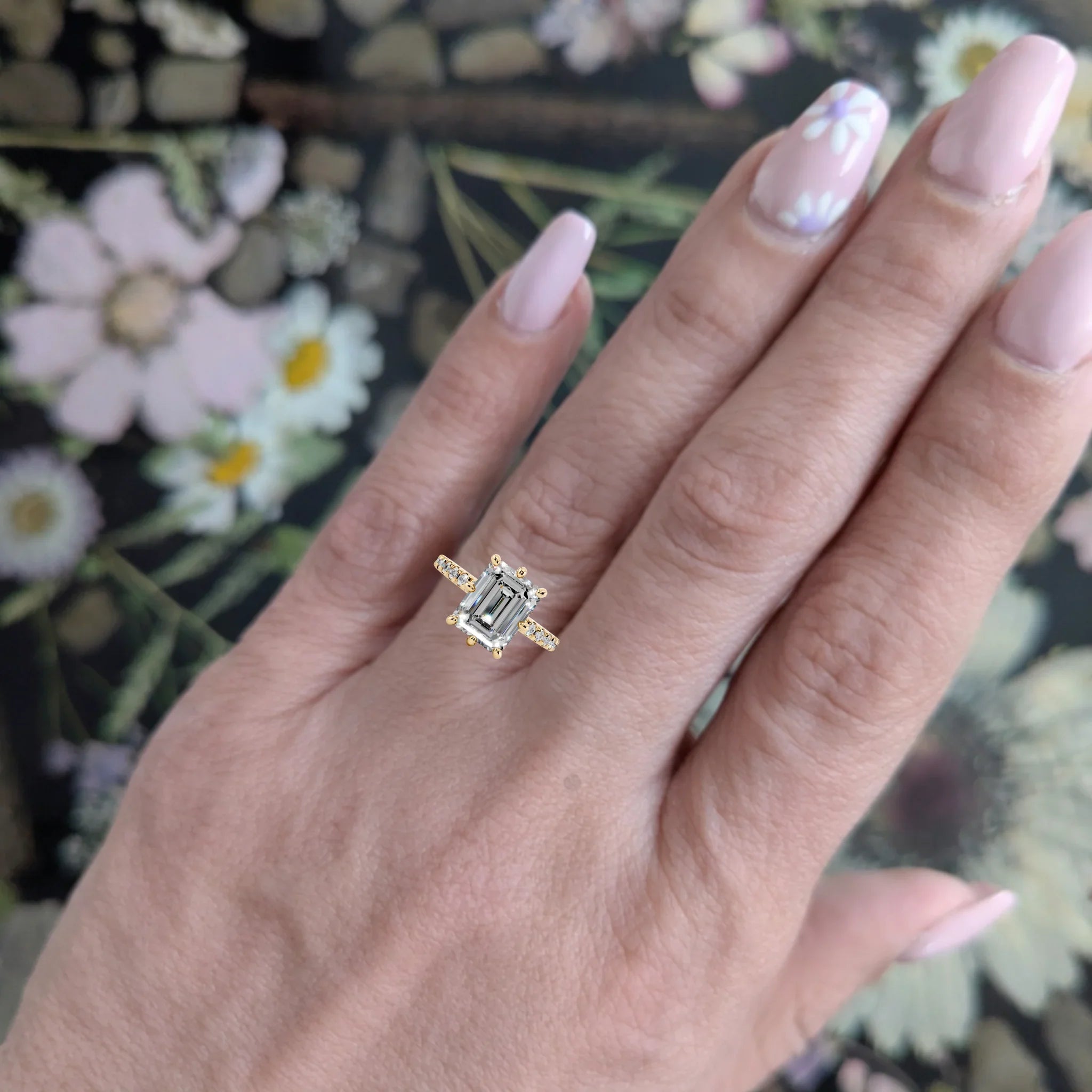 Emerald cut engagement ring with gold band on manicured hand against floral background