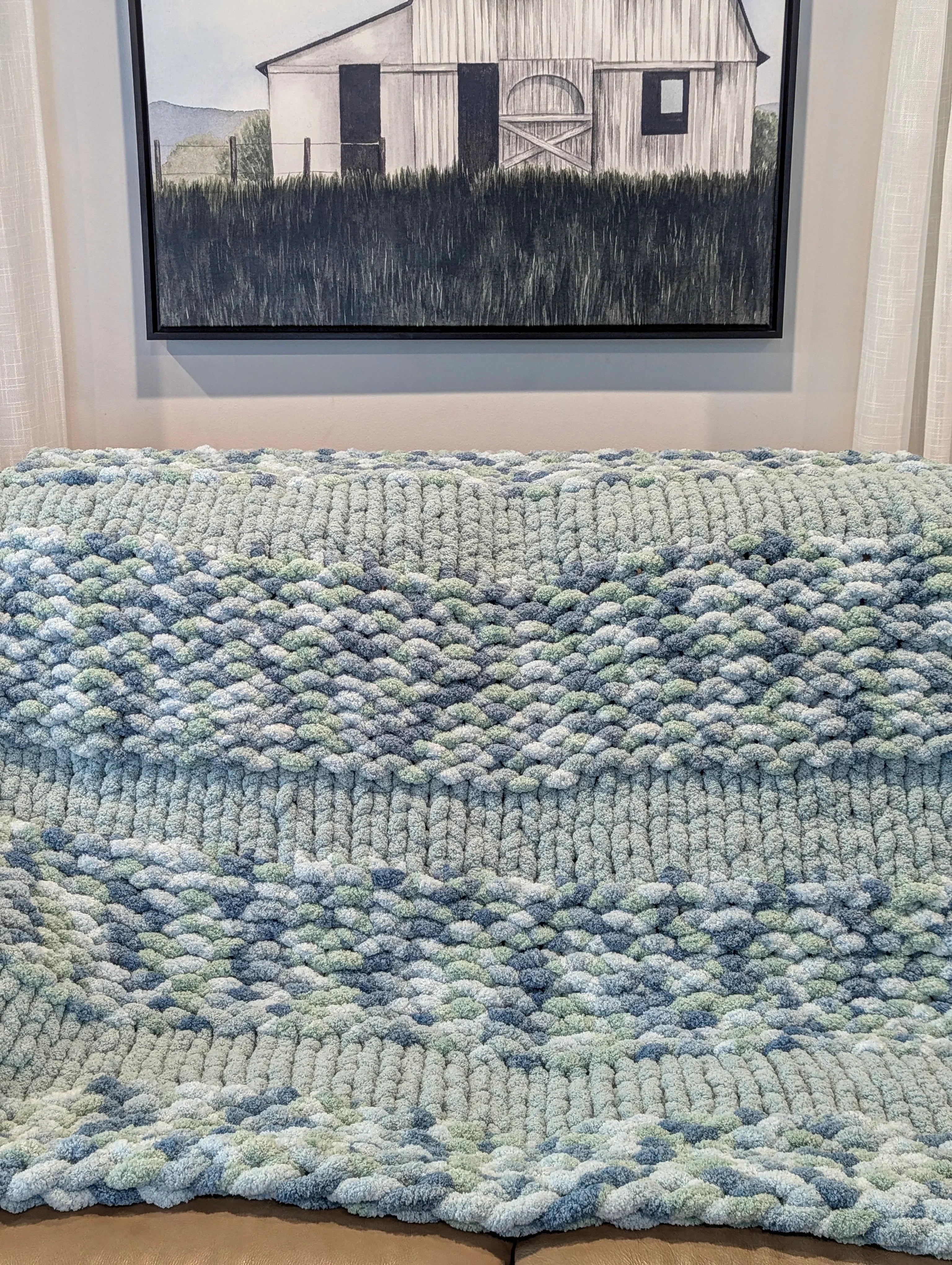 Chunky knit blue and green throw blanket on sofa, with farmhouse painting on wall.