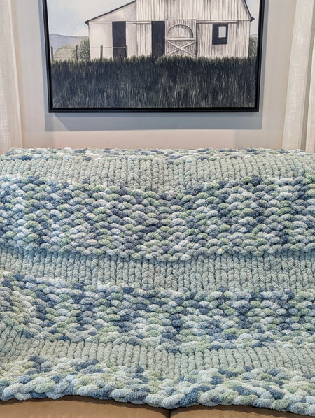 Chunky knit blue and green throw blanket on sofa, with farmhouse painting on wall.