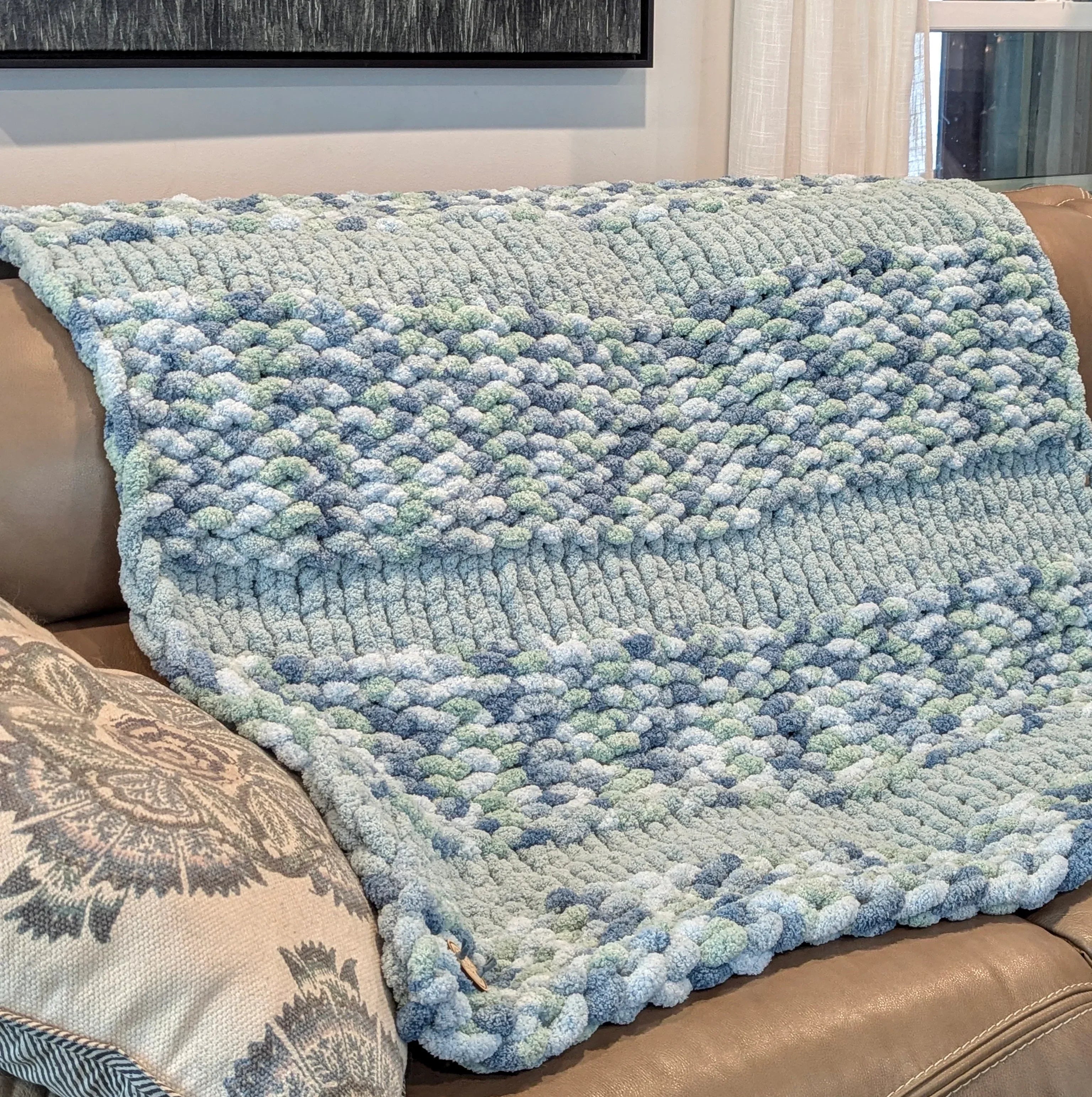 Chunky knit blue and green chenille throw blanket draped over a tan sofa in a living room.