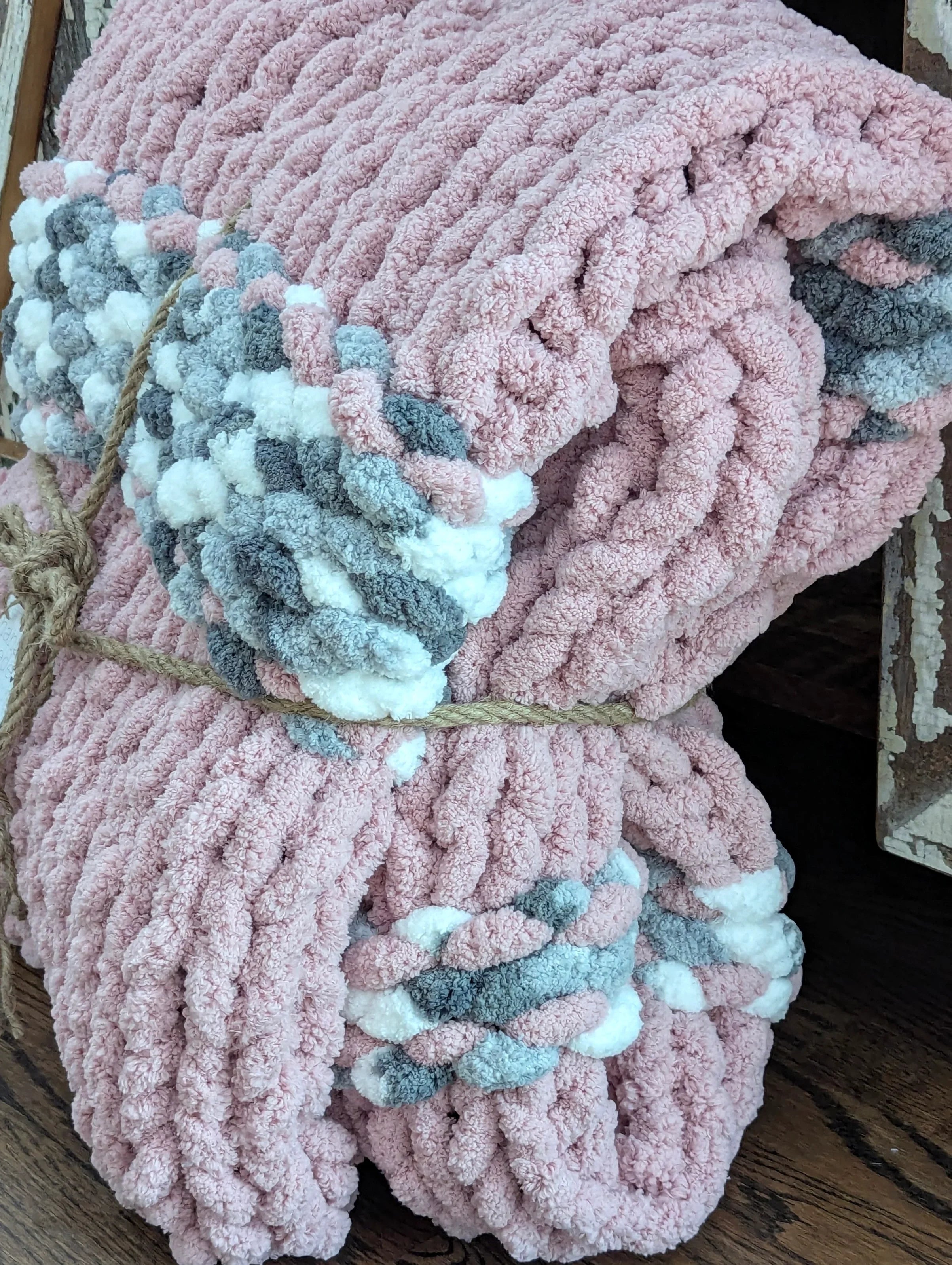 Chunky pink knitted blanket with braided gray and white accents, folded and tied with rope