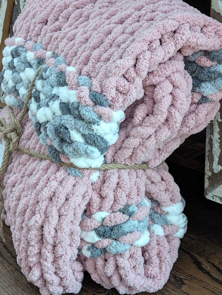 Chunky pink knitted blanket with braided gray and white accents, folded and tied with rope
