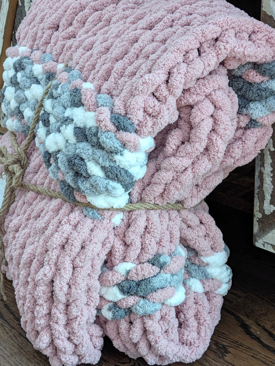 Chunky pink knitted blanket with braided gray and white accents, folded and tied with rope