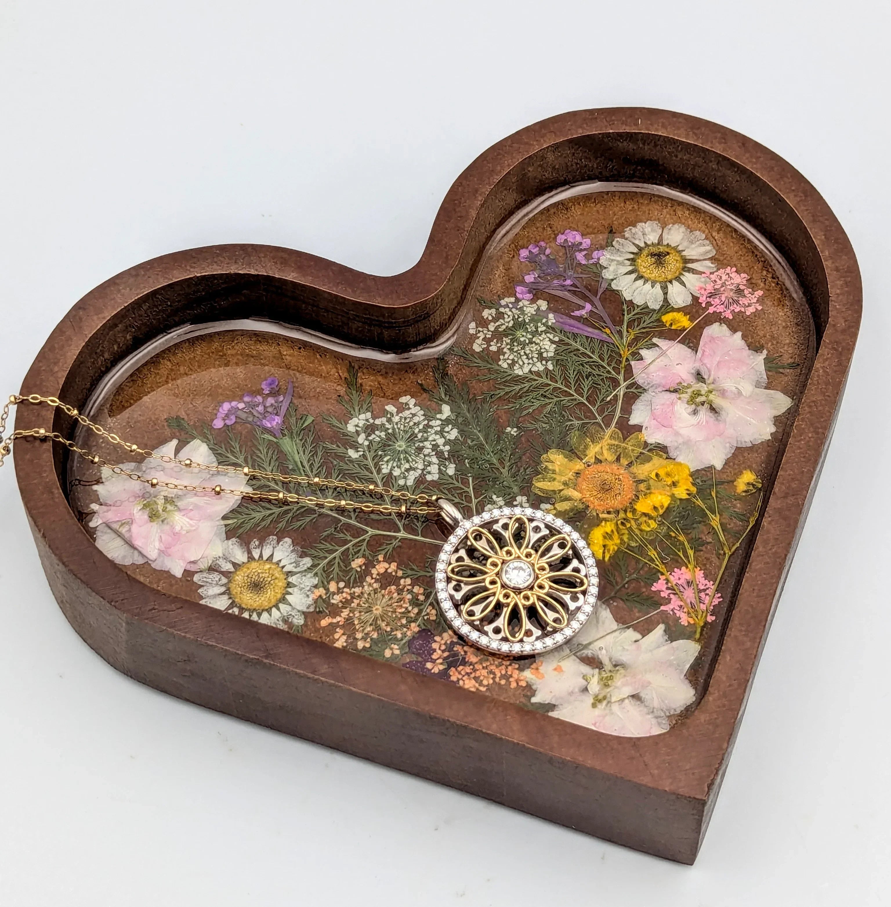 Gold floral pendant necklace on a heart-shaped wooden tray with pressed flowers