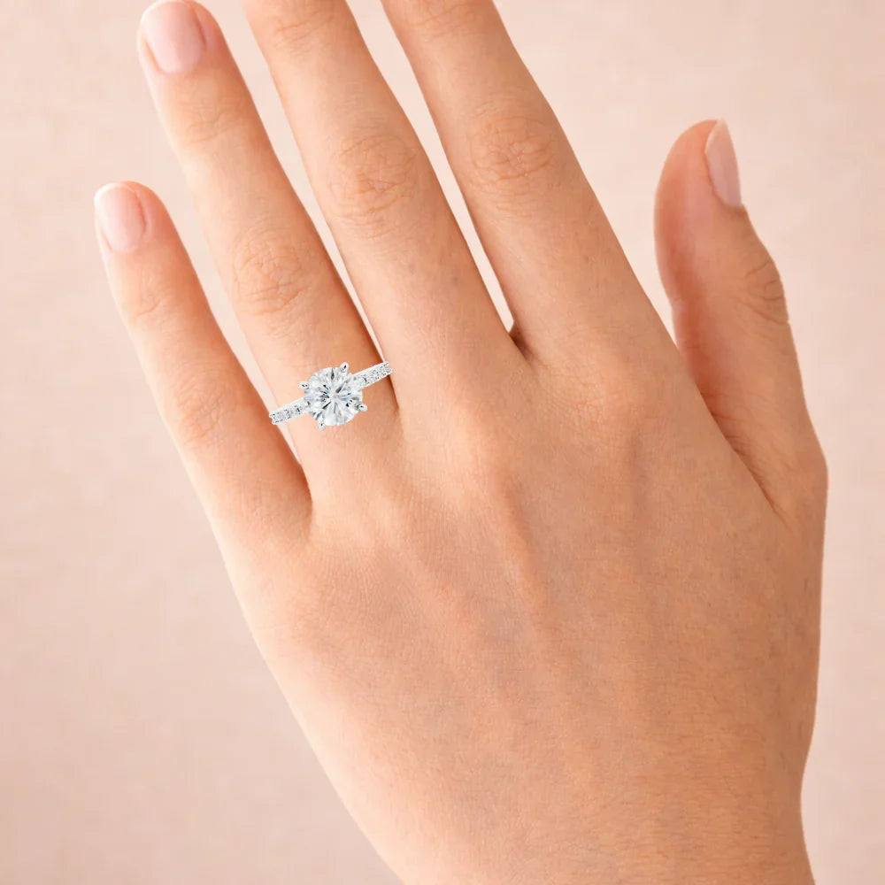 Hand wearing a round-cut diamond engagement ring with pavé band on a neutral background
