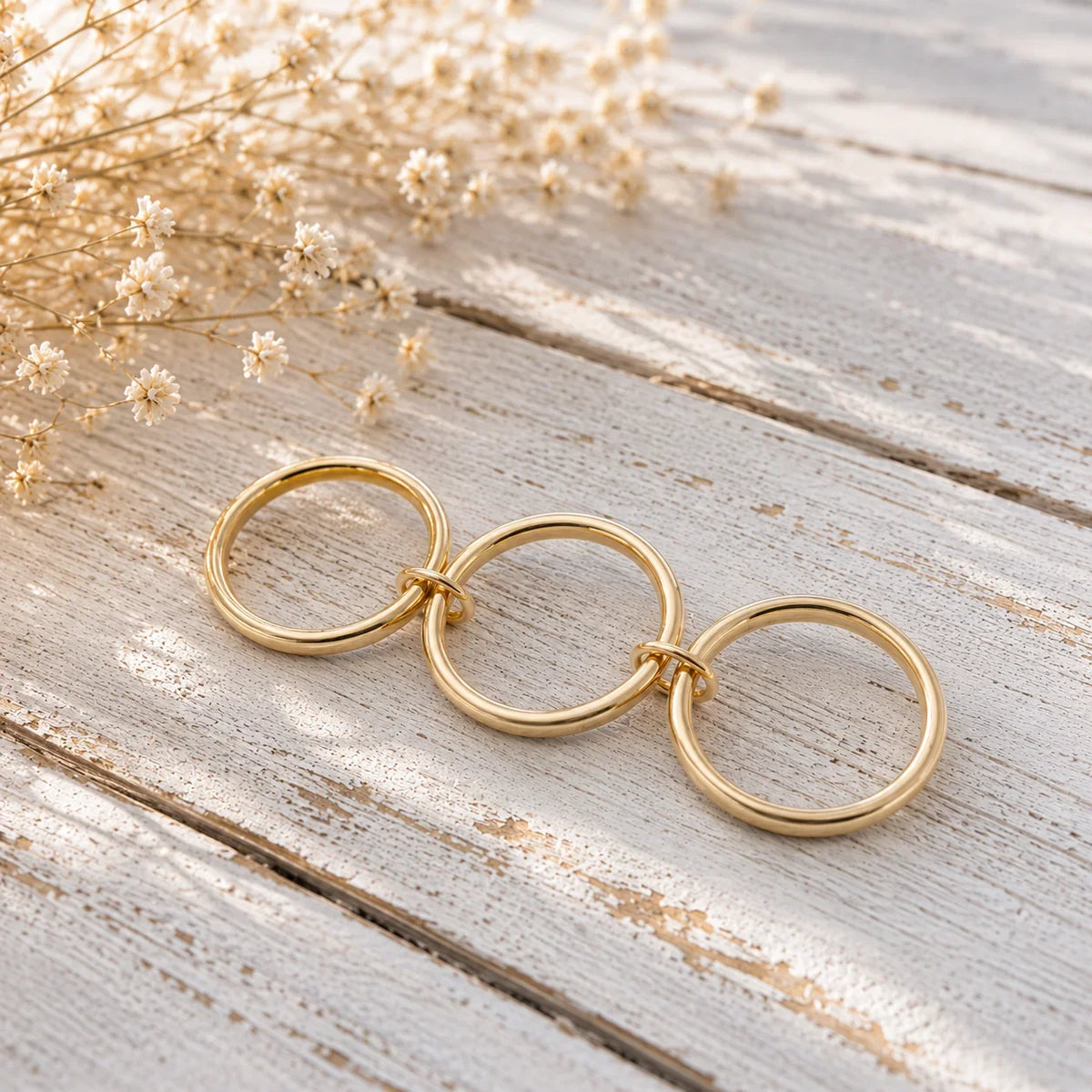 Three interlocking gold rings on rustic wood with dried flowers, symbolizing unity and custom jewelry design.